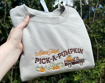 May include: A light gray sweatshirt with brown embroidered text that reads "Amber Farms Pick-A-Pumpkin". The text is accompanied by a brown truck with a pumpkin in the bed and three pumpkins on vines.