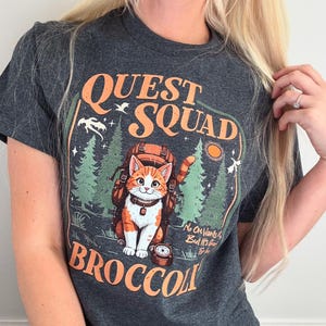Op de afbeelding: Donkergrijs t-shirt met een afbeelding van een kat met een rugzak, de woorden "QUEST SQUAD BROCCOLI" en boslandschap. De kat is oranje en wit. Het ontwerp bevat een kompas en een zon.