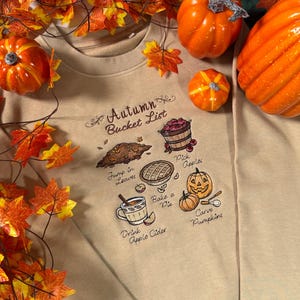 May include: A beige crewneck sweatshirt featuring an embroidered Autumn Bucket List design. The graphic includes illustrations of a leaf pile, a basket of apples, a pie, a mug of cider, and pumpkins with text reading Pick Apples, Jump in Leaves, Bake a Pie, Drink Apple Cider, and Carve Pumpkins.