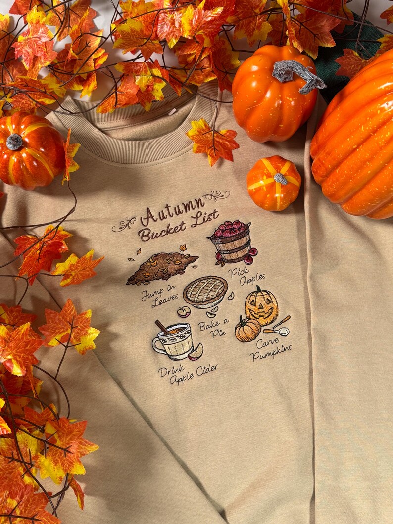 May include: A beige crewneck sweatshirt featuring an embroidered Autumn Bucket List design. The graphic includes illustrations of a leaf pile, a basket of apples, a pie, a mug of cider, and pumpkins with text reading Pick Apples, Jump in Leaves, Bake a Pie, Drink Apple Cider, and Carve Pumpkins.