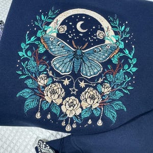 Puede incluir: Sudadera azul marino con bordado detallado. El dise&ntilde;o incluye una gran mariposa azul, una luna, estrellas y elementos florales. Decoraciones de mariposas adicionales est&aacute;n presentes.