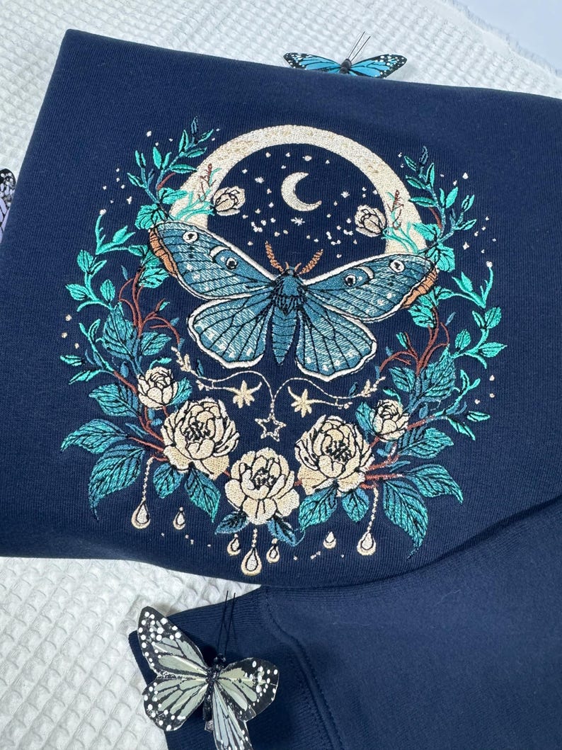 Puede incluir: Sudadera azul marino con bordado detallado. El dise&ntilde;o incluye una gran mariposa azul, una luna, estrellas y elementos florales. Decoraciones de mariposas adicionales est&aacute;n presentes.