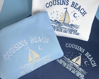Op de afbeelding: Drie sweaters met ronde hals in wit, lichtblauw en marineblauw. Elk exemplaar is voorzien van geborduurde tekst COUSINS BEACH, MASSACHUSETTS SUMMER UNDER THE MOON AND THE STARS, met een zeilboot, maansikkel en sterren.