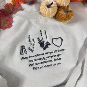 Könnte beinhalten: Ein beigefarbenes Sweatshirt mit Rundhalsausschnitt und schwarzer Stickerei eines Salzstreuers, eines Rosmarinzweigs, Lavendel und eines Herzens. Der Text lautet: Always throw spilled salt over your left shoulder. Keep rosemary by your garden gate. Plant roses and lavender, for luck. Fall in love whenever you can.
