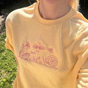 Può includere: Una t-shirt giallo pallido con un ricamo a linee rosa scuro sul petto. Il ricamo raffigura una rana, diversi funghi di varie dimensioni, una piccola ape in volo e una lumaca con un guscio a spirale dettagliato sul dorso.