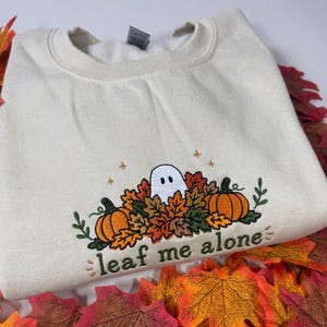 Pode incluir: Uma camisola de gola redonda cor de creme com um design bordado de um fantasma branco a espreitar de um monte de folhas de outono coloridas e duas abóboras cor de laranja. O texto "leaf me alone" está bordado a verde sob a ilustração.