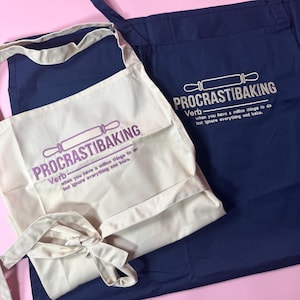 Può includere: Un grembiule color crema e un grembiule blu navy, entrambi con un'icona di un mattarello ricamata e il testo "PROCRASTIBAKING Verb: when you have a million things to do but ignore everything and bake." Il grembiule color crema ha il testo viola.