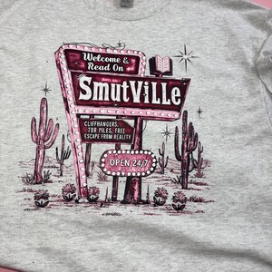 May include: A light gray t-shirt featuring a retro-style sign that reads "SmutVille." The sign is pink and white, with the words "Welcome & Read On" above and "Open 24/7" below. The design includes cacti and a book.