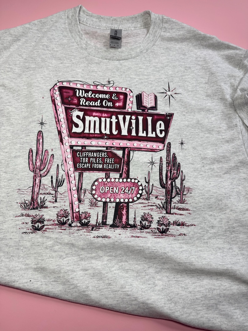 May include: A light gray t-shirt featuring a retro-style sign that reads "SmutVille." The sign is pink and white, with the words "Welcome & Read On" above and "Open 24/7" below. The design includes cacti and a book.