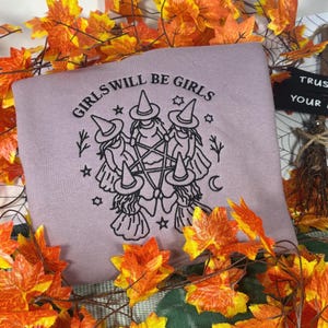 Op de afbeelding: Een mauve sweatshirt met zwarte borduurwerk van vijf figuren met puntmutsen, gerangschikt in een cirkel rond een centraal pentagram. De tekst "GIRLS WILL BE GIRLS" staat in een boog boven de figuren, geaccentueerd door kleine sterren.
