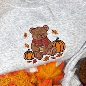 May include: A light grey crewneck sweatshirt featuring an embroidered design of a brown teddy bear wearing a red scarf and holding a coffee cup, surrounded by two orange pumpkins and several scattered autumn leaves in shades of orange and red.