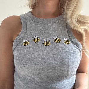 May include: A grey vest top featuring five embroidered cartoon bees in yellow and black with white details. The vest is sleeveless and has a ribbed texture. The bees are arranged in a horizontal row.
