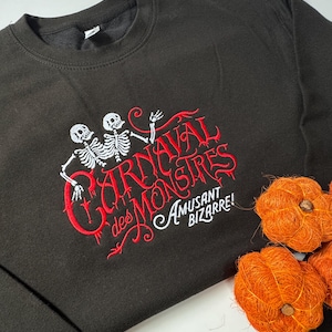 Peut inclure: Un sweat-shirt noir à col rond avec une broderie représentant deux squelettes blancs. Le texte rouge indique "CARNAVAL des MONSTRES" et le texte blanc en dessous précise "AMUSANT BIZARRE!" Le motif inclut des volutes et ornements rouges.