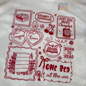 May include: Cream-colored t-shirt with a red graphic design featuring book-themed illustrations and text. The design includes phrases like "Enemies to Lovers," "Just One More Chapter," and "Born to Read."