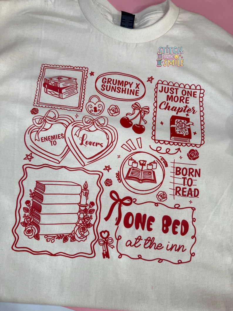 May include: Cream-colored t-shirt with a red graphic design featuring book-themed illustrations and text. The design includes phrases like "Enemies to Lovers," "Just One More Chapter," and "Born to Read."