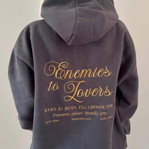 Può includere: Felpa con cappuccio grigio scuro con testo dorato "Enemies to Lovers". Il design include le frasi "Even in ruin, I'll choose you" e "Favourite colour? Morally Grey."