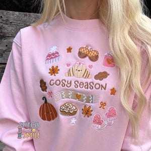May include: Pale pink sweatshirt with embroidered autumnal motifs, such as a croissant, acorns, leaves, and the words "COSY SEASON". The design also includes a pumpkin, mittens, and a scarf.