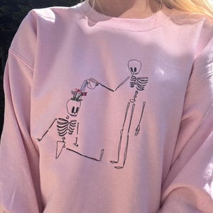 Könnte beinhalten: Ein hellrosa Sweatshirt mit einer schwarzen Strichzeichnung von zwei Skeletten. Ein Skelett gießt Blumen, die aus dem Kopf des anderen wachsen. Das Sweatshirt hat eine einfarbige, hellrosa Farbe.