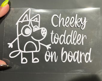 May include: White vinyl decal featuring a cartoon dog character with the text "Cheeky toddler on board". The decal is on a clear background, suitable for application on a car window or similar surface.