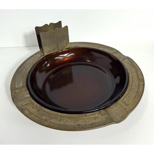 May include: Vintage ashtray with a brown glass bowl and a brass-colored metal rim. The rim has a decorative pattern and a built-in cigarette holder. The ashtray is designed to hold cigarettes and catch ashes.
