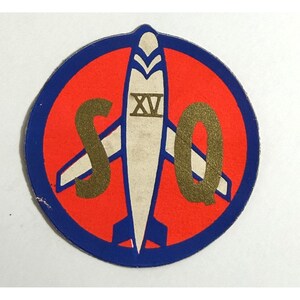 May include: A vintage patch featuring a red circle with a blue border. Inside, a white rocket shape is centered, with the Roman numeral XV. Gold letters "SQ" flank the rocket. A collectible item.
