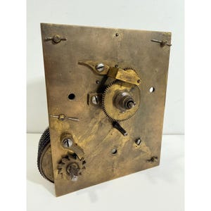 May include: A rectangular brass clock movement plate featuring a central gear, a ratchet wheel at the bottom, and various metal screws and pins. The surface shows a weathered, aged patina with visible mechanical components attached to the front.