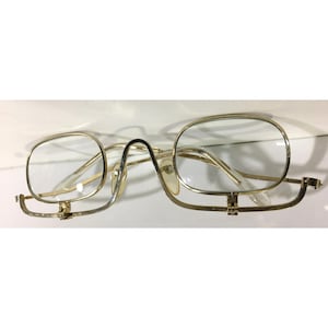 May include: Vintage gold-tone eyeglasses with clear lenses. The frames have a rectangular shape with a bridge and folding side arms. The glasses are designed for vision correction and are likely from a previous era.