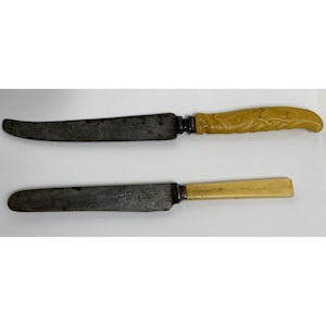 May include: Two antique knives with dark, aged blades. One knife has a curved blade and a carved, yellow handle. The other knife has a straight blade and a light yellow handle. Both knives show signs of wear.