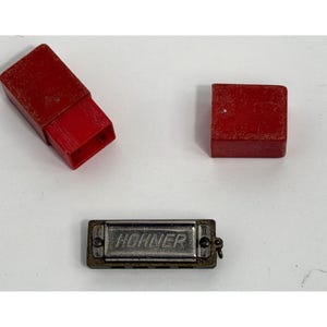 May include: A Hohner harmonica with a silver metal body and the brand name in the center. The harmonica is accompanied by two red cases, one rectangular and one square, likely for storage and protection. The items are on a white surface.