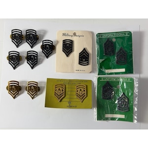 May include: A collection of military rank insignia pins in black and gold tones. Some pins are loose, while others are attached to cream, green, or yellow cardboard backing cards labeled Military Insignia or Uniform Insignia, with some in plastic.