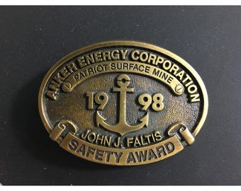May include: Oval-shaped bronze award with the text "ANKER ENERGY CORPORATION PATRIOT SURFACE MINE" and "1998" with an anchor symbol. The text "JOHN J. FALTIS SAFETY AWARD" is also present. The award has a vintage appearance.