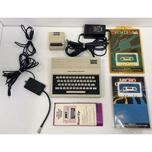 May include: A beige Radio Shack TRS-80 Micro Color Computer with a black keyboard, power adapter, RF modulator, and a small peripheral device. Includes a purple Start-Up manual and two packaged software cassettes labeled Math Design and Micro Color.