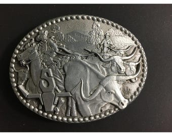 May include: Oval silver-tone belt buckle with a Western theme. The buckle features a detailed relief of a cowboy on horseback, cattle, and the letters "S V". The border is adorned with small, raised dots.
