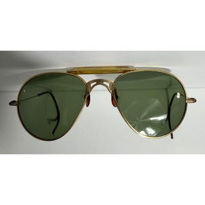 May include: Gold-framed aviator sunglasses with green lenses. The sunglasses feature a double bridge and a curved brow bar. The lenses are teardrop-shaped, and the frames have a classic, vintage design. The temples are thin and extend to the ear pieces.