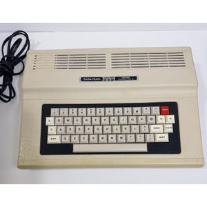 May include: A beige Radio Shack TRS-80 Color Computer 2 with a QWERTY keyboard, a black keyboard base, and a red break key. The unit features a top ventilation grille and a black power cord coiled to the left. The label displays the text Radio Shack TRS-80 Color Computer 2.