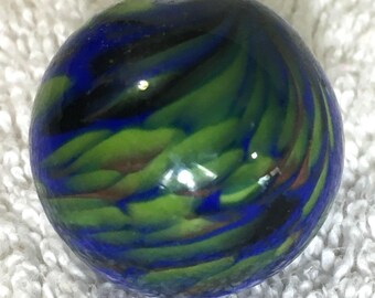 May include: A glass marble with a swirling pattern of blue, green, and brown colors. The marble is round and has a glossy finish. The design resembles a miniature galaxy or a stylized landscape.