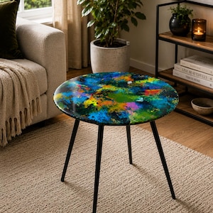 Può includere: Un tavolino rotondo con un design astratto colorato. Il piano del tavolo presenta un vivace mix di tonalità blu, verdi, arancioni e gialle. Ha una struttura in metallo nero con tre gambe. Il diametro del tavolo è di circa 60 cm.