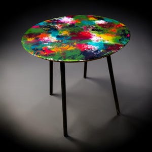 Può includere: Un tavolino rotondo con un piano dipinto astratto e colorato. Il piano presenta un vivace mix di tonalità verdi, rosse, blu e gialle. Il tavolo ha una struttura e gambe in metallo nero.