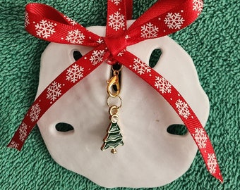 May include: A white seashell ornament with a red ribbon with white snowflakes printed on it. A gold charm with a green Christmas tree hangs from the ribbon.