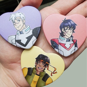 May include: Three heart-shaped buttons with cartoon characters. The first button is purple with a white-haired character wearing a white and black uniform. The second button is pink with a blue-haired character wearing a red and white uniform. The third button is yellow with a brown-haired character wearing a yellow and green uniform.