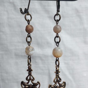 May include: A pair of dangle earrings with a bronze finish. Each earring features a decorative bee charm, linked to a chain with three stone beads. The earrings are suspended from a bronze ear wire.