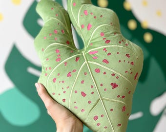 Può includere: Un cuscino a forma di foglia, in morbido tessuto verde brillante, decorato con macchie rosa e venature ricamate. Il cuscino è tenuto davanti a un muro con un design di foglie tropicali.