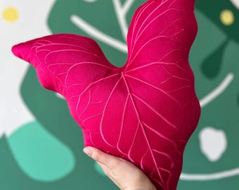 Può includere: Un cuscino a forma di foglia fucsia vibrante con ricami di venature dettagliati. Il cuscino è tenuto contro uno sfondo di parete verde e bianca con un motivo a foglie. Un oggetto decorativo.