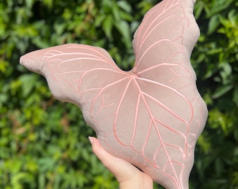 Può includere: Un cuscino a forma di foglia rosa pallido con dettagli ricamati. Il cuscino è tenuto in mano su uno sfondo di fogliame verde. Il design della foglia presenta cuciture rosa più chiare, conferendo un aspetto morbido e strutturato.