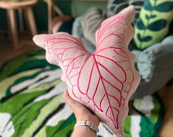 Può includere: Un cuscino in velluto rosa con un motivo a foglia rosa ricamato. Il cuscino ha la forma di una foglia e ha un contorno rosa.