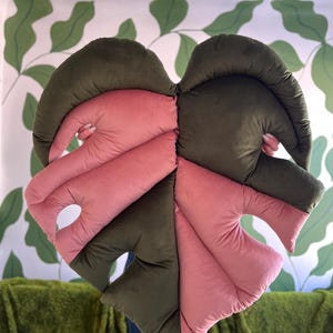 Puede incluir: Un gran cojín con forma de hoja, con un diseño bicolor. Una mitad es verde oliva oscuro, la otra rosa suave. El cojín tiene una textura acolchada y un agujero cerca del centro, que se asemeja a una hoja de Monstera. El fondo presenta un patrón de hojas verdes.