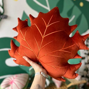 May include: A large, rust-coloured leaf-shaped cushion with detailed vein embroidery. The plush cushion is held up against a green and white patterned wall. The cushion is a decorative accent piece.