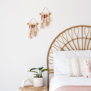 Two rainbow macrame wall hangings hanged above a bed in a kids bedroom.