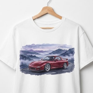 Pode incluir: Camiseta branca com uma ilustração em aquarela de um carro esportivo vermelho em uma estrada de montanha. O carro é o ponto focal, com um design detalhado. O fundo mostra uma vista panorâmica das montanhas e um céu nublado.