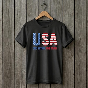 Peut inclure: Un t-shirt noir &agrave; manches courtes avec le texte USA dans un motif de drapeau am&eacute;ricain rouge, blanc et bleu. Sous les grandes lettres, le texte indique ONE NATION. ONE TEAM. en bleu et rouge. Le t-shirt est sur un cintre en bois.